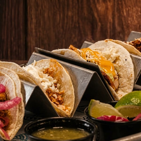 A metal tray holds five tacos in stands, with various fillings, sauces, and lime wedges.