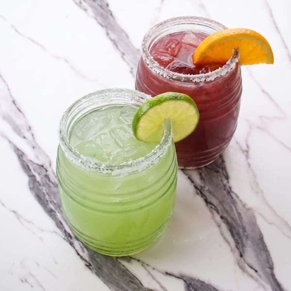 Green and red cocktails with salt rims, garnished with lime and orange, on marble.