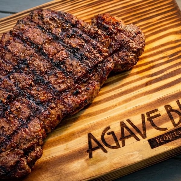 Grilled steak with grill marks on a wooden cutting board with the Agave Blue logo.