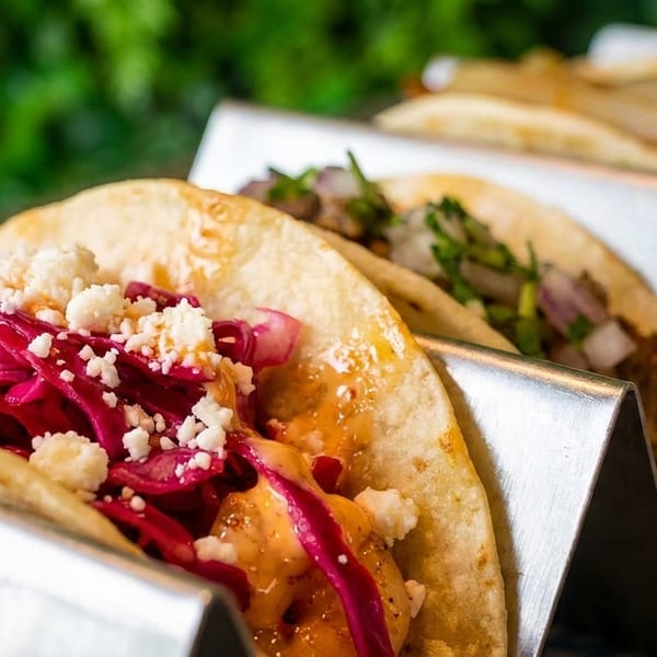 Close-up of four tacos in metal holders, with red cabbage and cheese on the front taco.