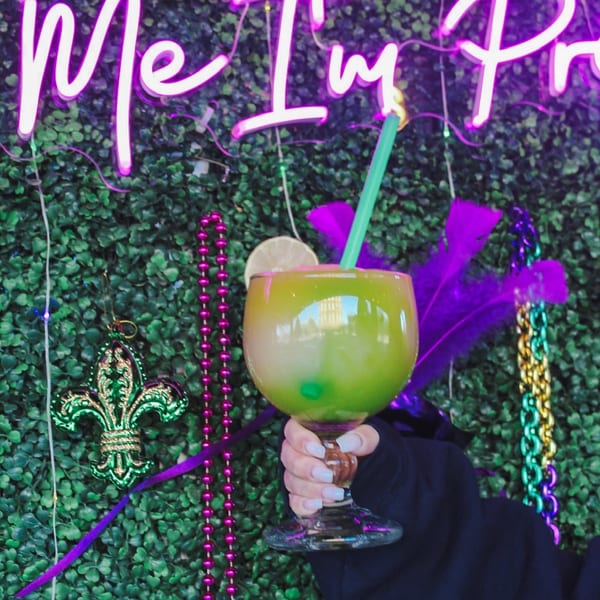Hand holding a green cocktail with a lime slice and straw against a neon sign and Mardi Gras decor.