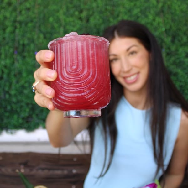 Smiling woman holding up a vibrant red cocktail in a textured glass against a green hedge.