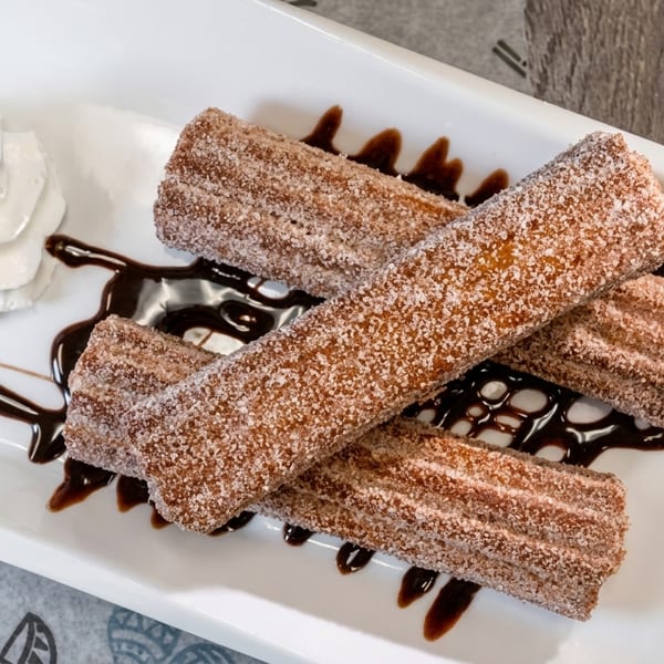 Four sugar-dusted churros with chocolate drizzle and a dollop of whipped cream on a white plate.