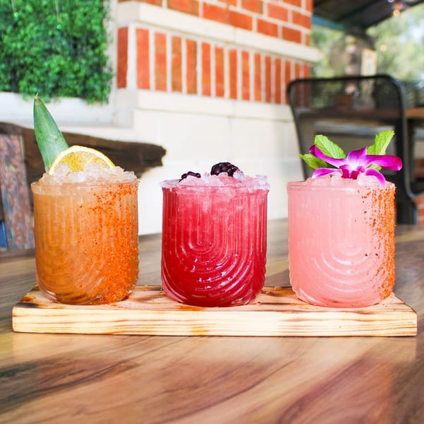 Three colorful cocktails with ice and garnishes on a wooden board, with a brick wall background.