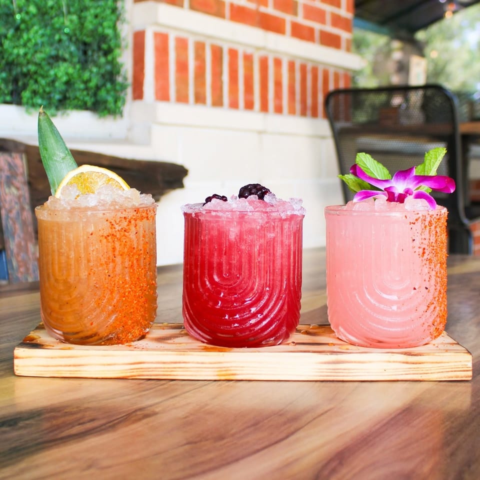 Three colorful cocktails with ice and garnishes on a wooden board, with a brick wall background.