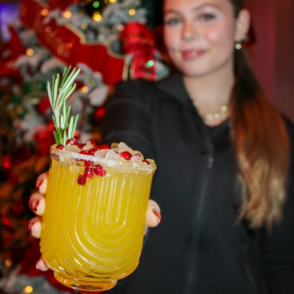 A woman holds a festive yellow cocktail garnished with rosemary and pomegranate seeds in front of a Christmas tree.