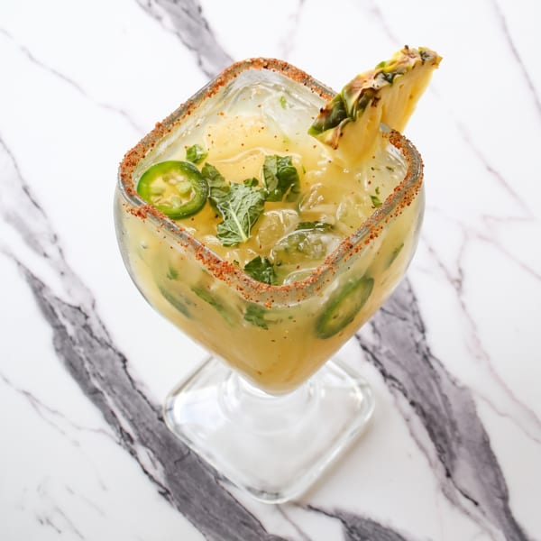 Spicy pineapple margarita with jalapeño slices, mint, and a chili-lime rim on a marble surface.