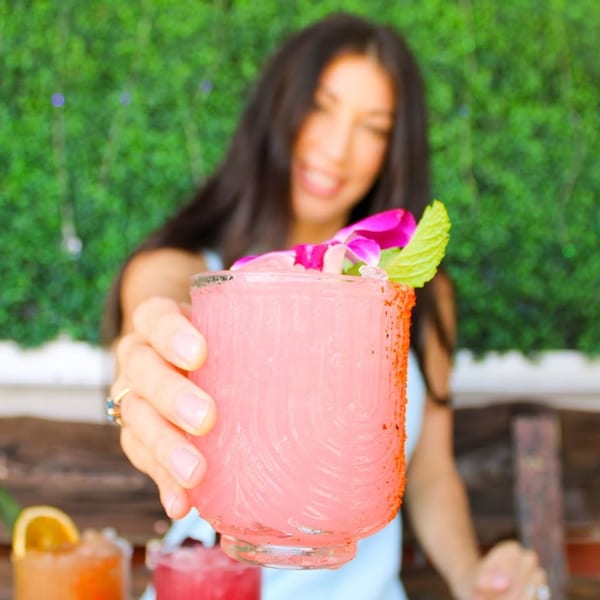 A woman holds out a pink cocktail garnished with a purple orchid and mint leaf.