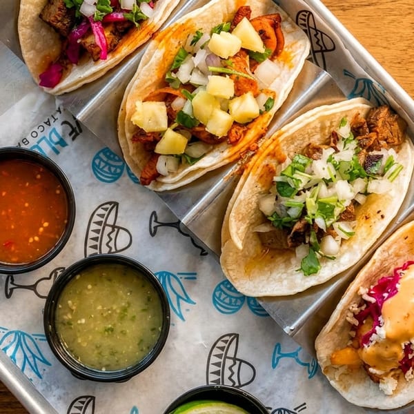 A metal tray holds five diverse tacos, two salsa bowls, and a lime wedge on branded paper.